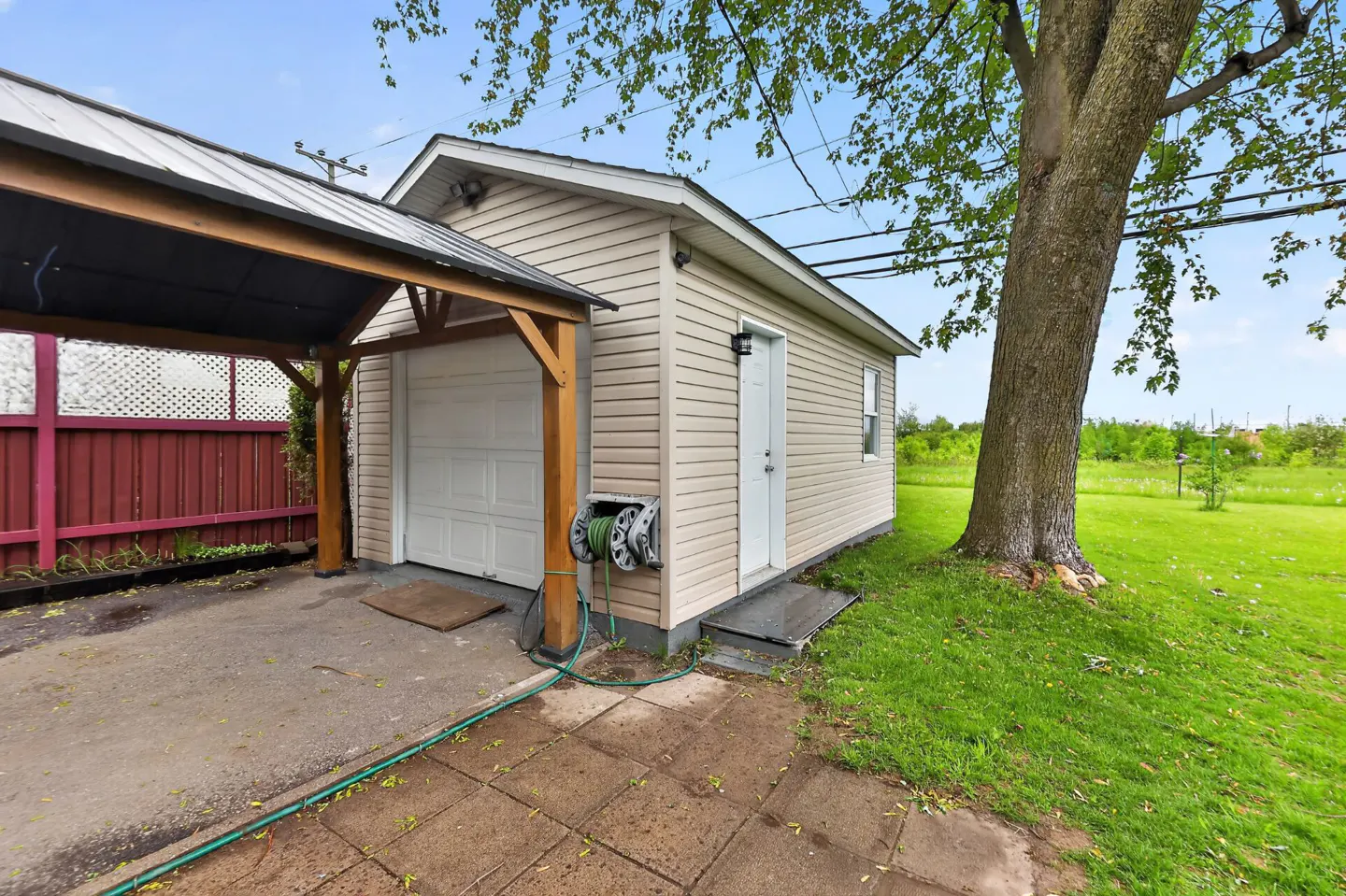 Tan garage with white door and side entrance, attached to a carport with a brown frame. Green lawn and tree to the right.