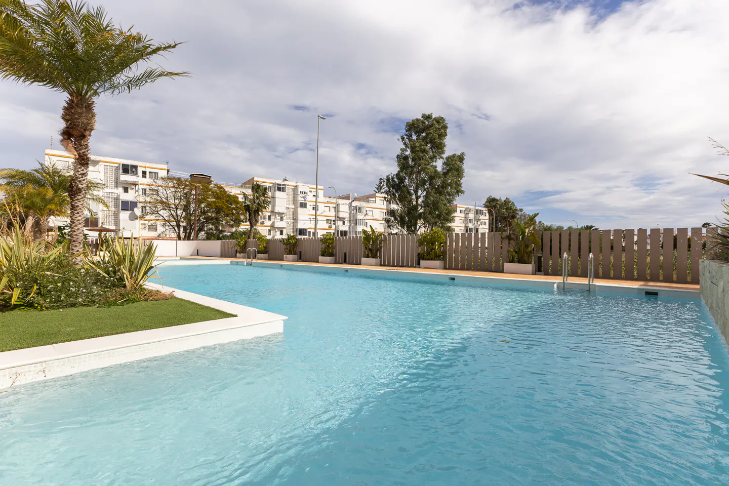 Outdoor pool with light blue water, surrounded by a white deck, palm trees, and a white apartment building under a cloudy sky.