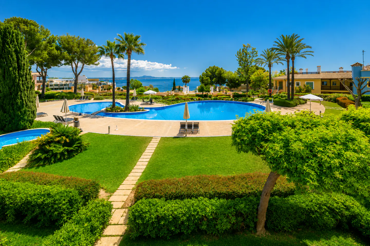 A large, blue, kidney-shaped swimming pool surrounded by lush green lawns, palm trees, and a view of the Mediterranean Sea.