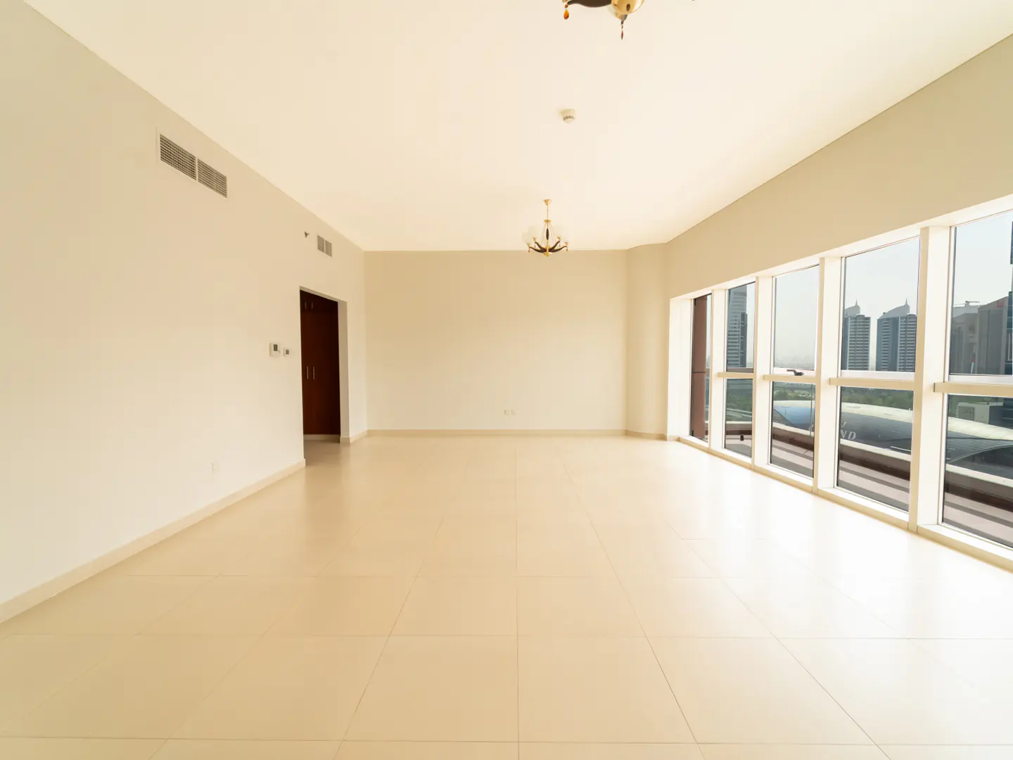 Spacious, empty room with beige tiled floors, white walls, modern chandeliers, and large windows overlooking city towers.