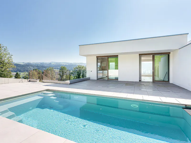 A modern white house with a blue swimming pool and a view of green hills under a clear blue sky.