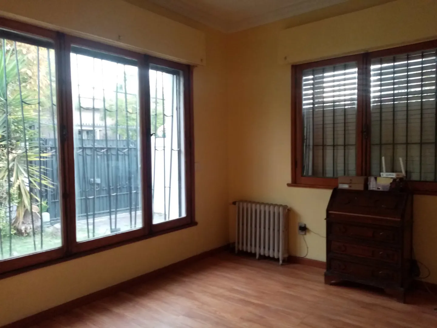 A room with wood floors, yellow walls, and two windows. A radiator sits under one window, and a wooden desk is near the other.