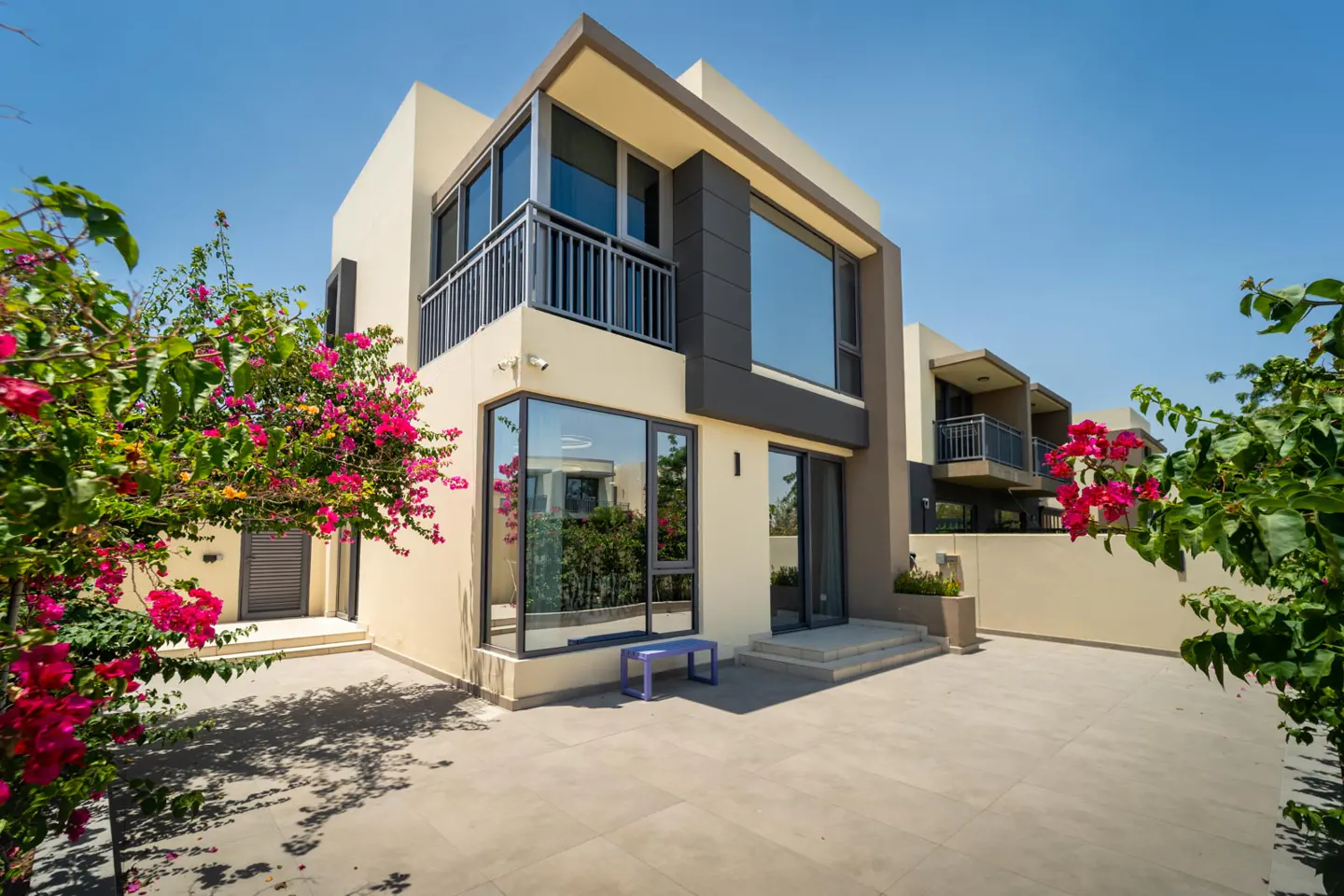 Modern two-story home with beige walls, gray accents, large windows, and a balcony. Pink bougainvillea frames the patio.