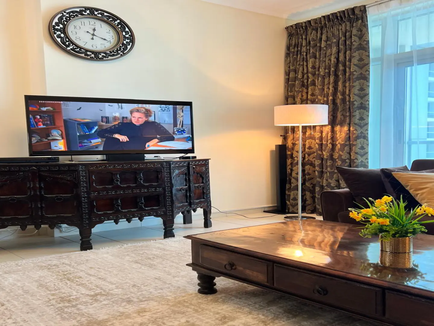 Living room with dark wood furniture, a large TV, and a cream-colored rug. A clock hangs above the TV. Yellow flowers sit on the coffee table.