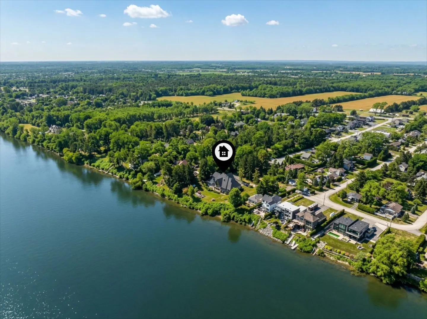 Aerial view of waterfront homes on a sunny day, with a house icon marking a specific property. Trees and fields in the background.