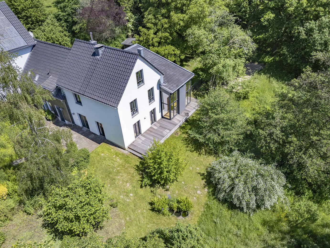 Aerial view of a white two-story house with a dark tiled roof, wooden deck, and lush green garden surrounded by trees.