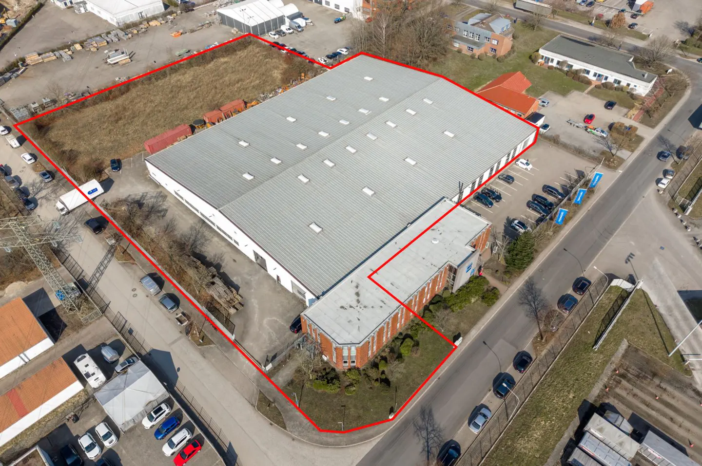 Aerial view of a large, gray warehouse with a red outline, surrounded by parking, roads, and a vacant lot.