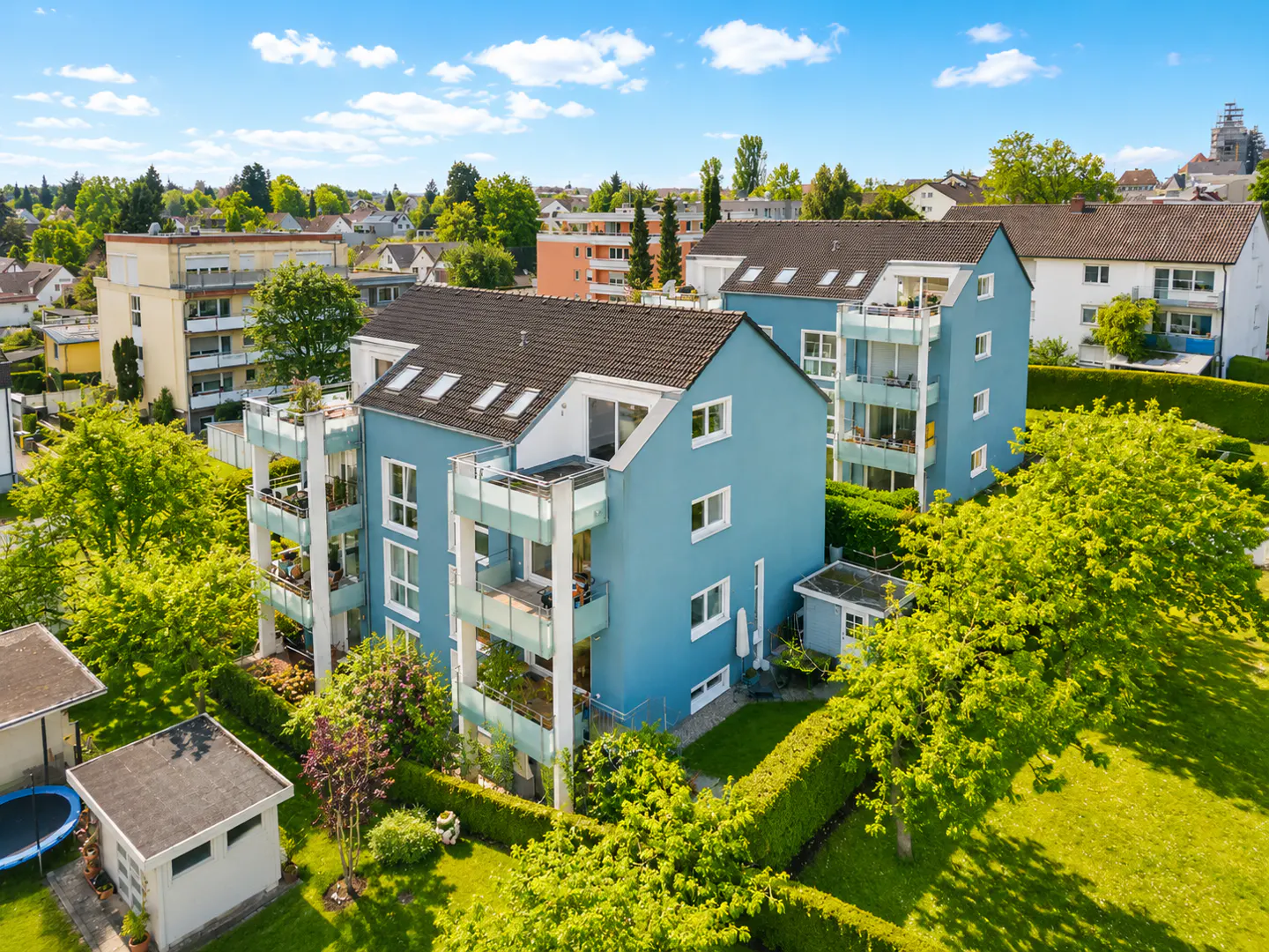 Aerial view of a blue multi-story apartment building with balconies, surrounded by green trees and a lawn on a sunny day.