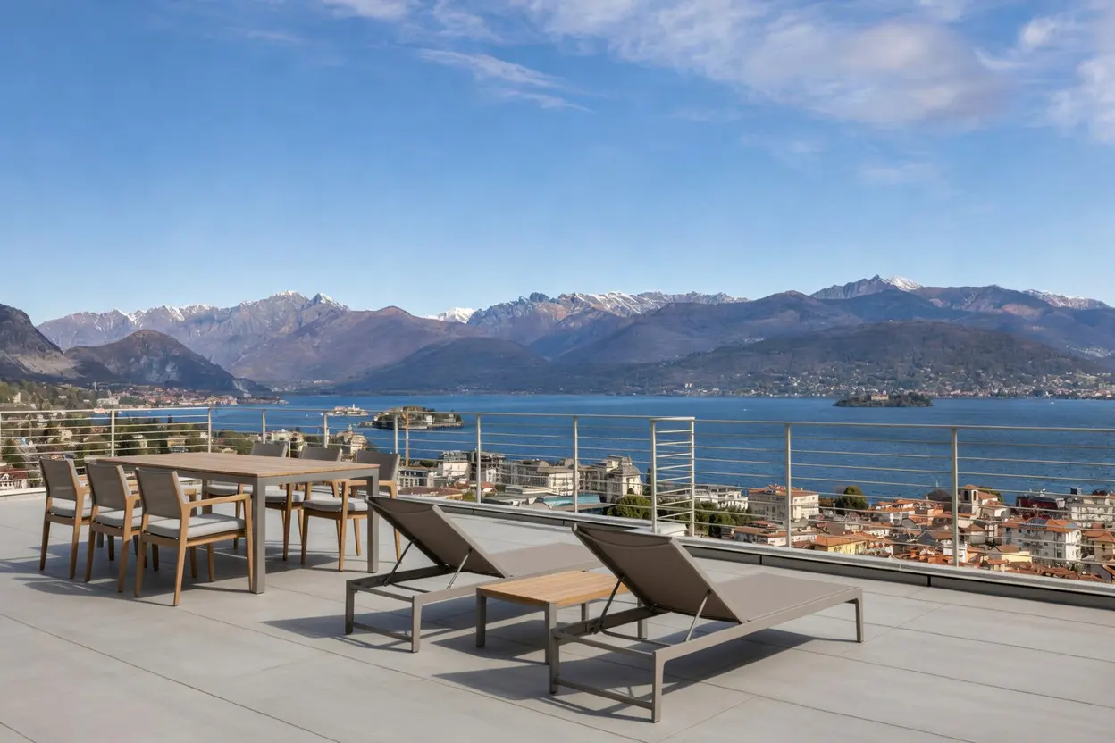 Rooftop patio with dining table, chairs, and lounge chairs overlooking a lake, town, and snow-capped mountains under a blue sky.