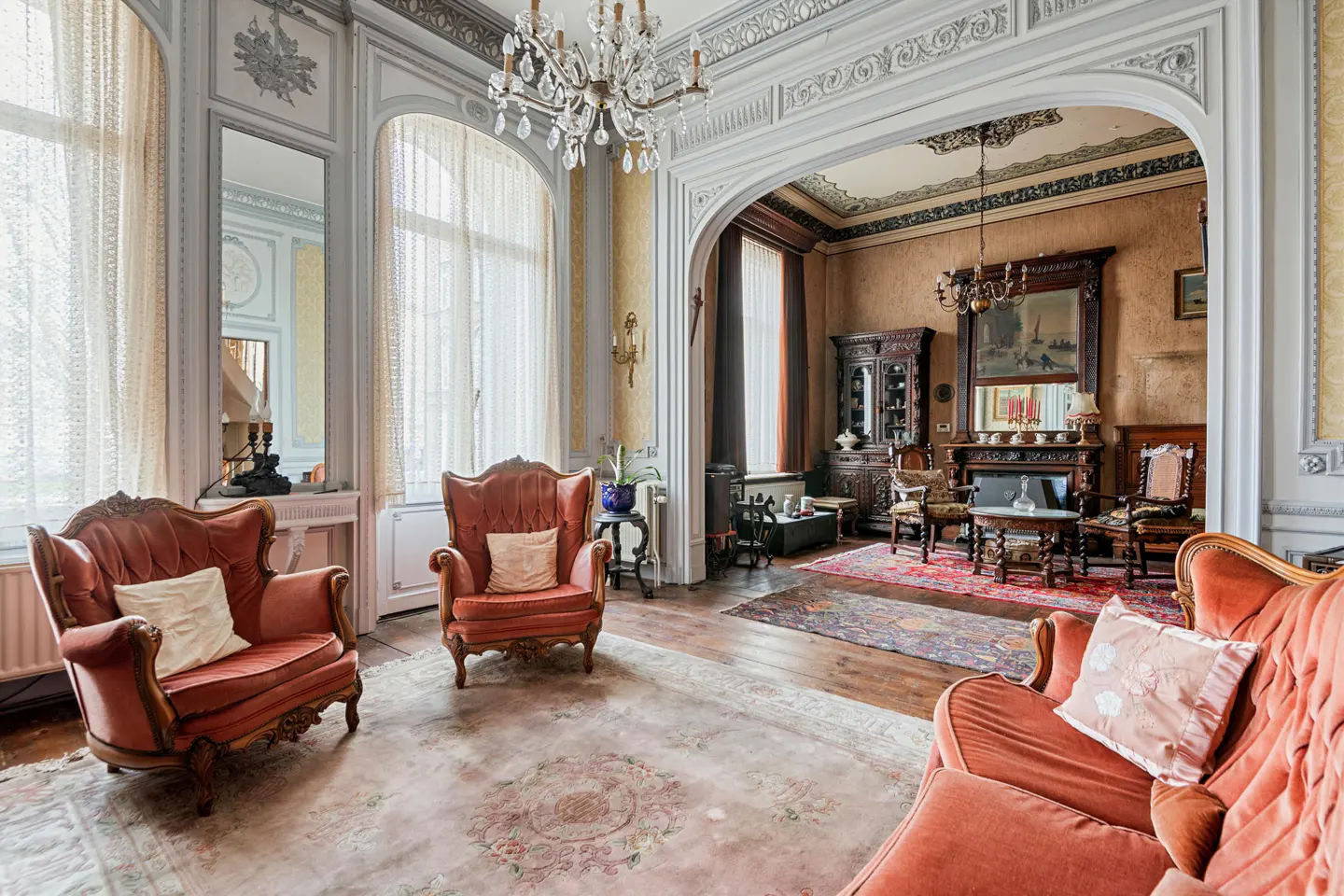 Ornate living room with red armchairs, a crystal chandelier, and an arched doorway leading to a second room with a fireplace.