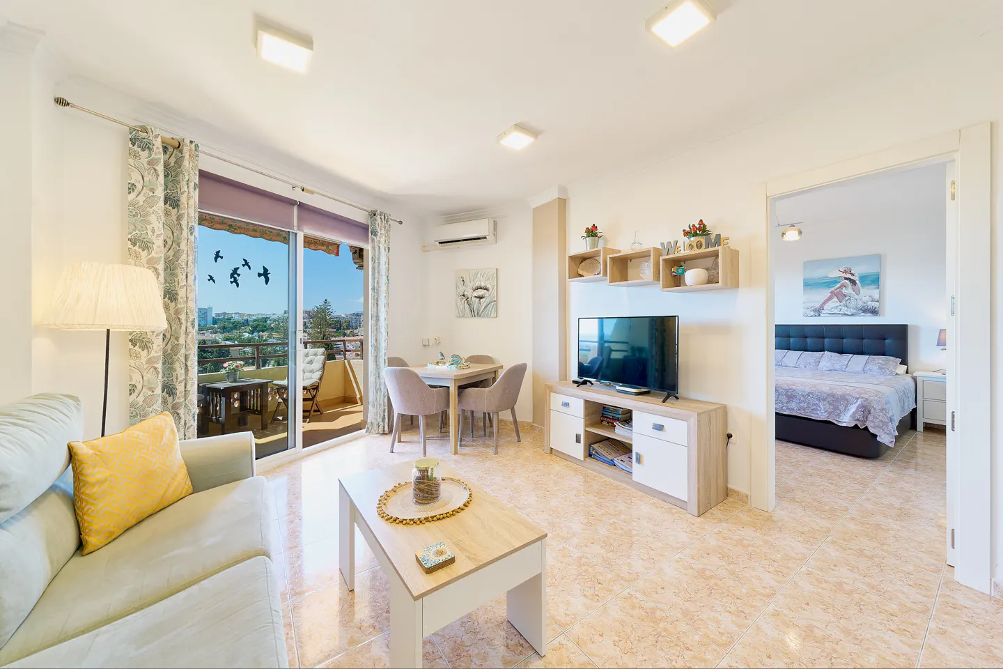 Bright living room with a sofa, TV, dining table, and balcony with a city view. Bedroom visible through doorway.