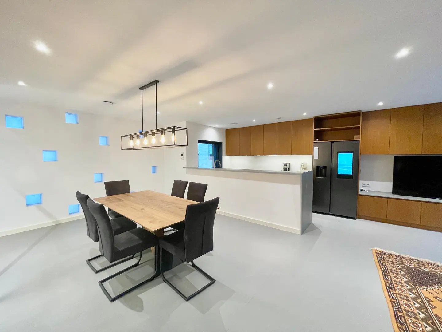 A modern, open-concept dining and kitchen area with a wooden table, six gray chairs, and a rectangular light fixture. The kitchen features wood cabinets and a black refrigerator.