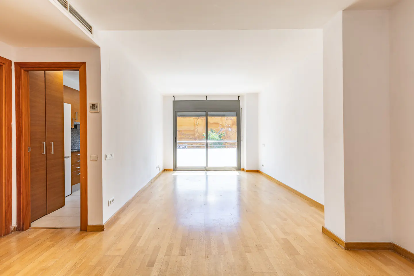 Empty room with light wood floors, white walls, and a sliding glass door to a balcony. A doorway leads to a kitchen.