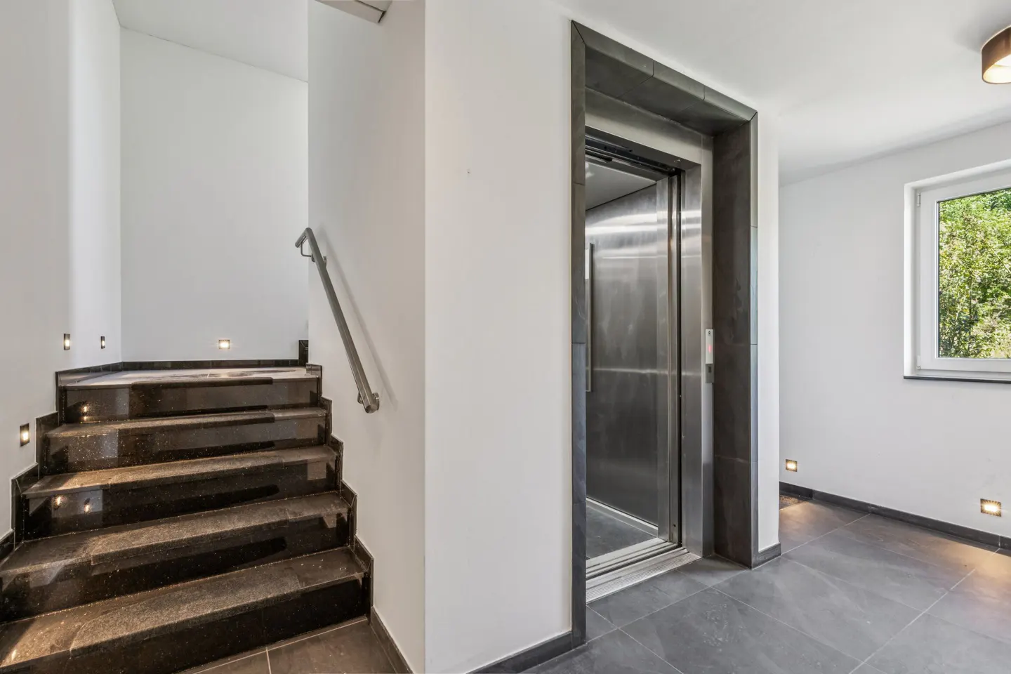 Interior view of a modern home with black marble stairs, a silver elevator, and a window.
