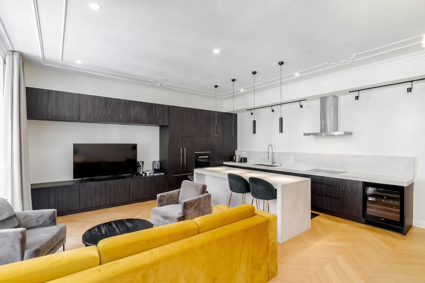 Modern open-plan living area with a yellow sofa, grey armchairs, and a sleek kitchen featuring dark cabinets and an island.