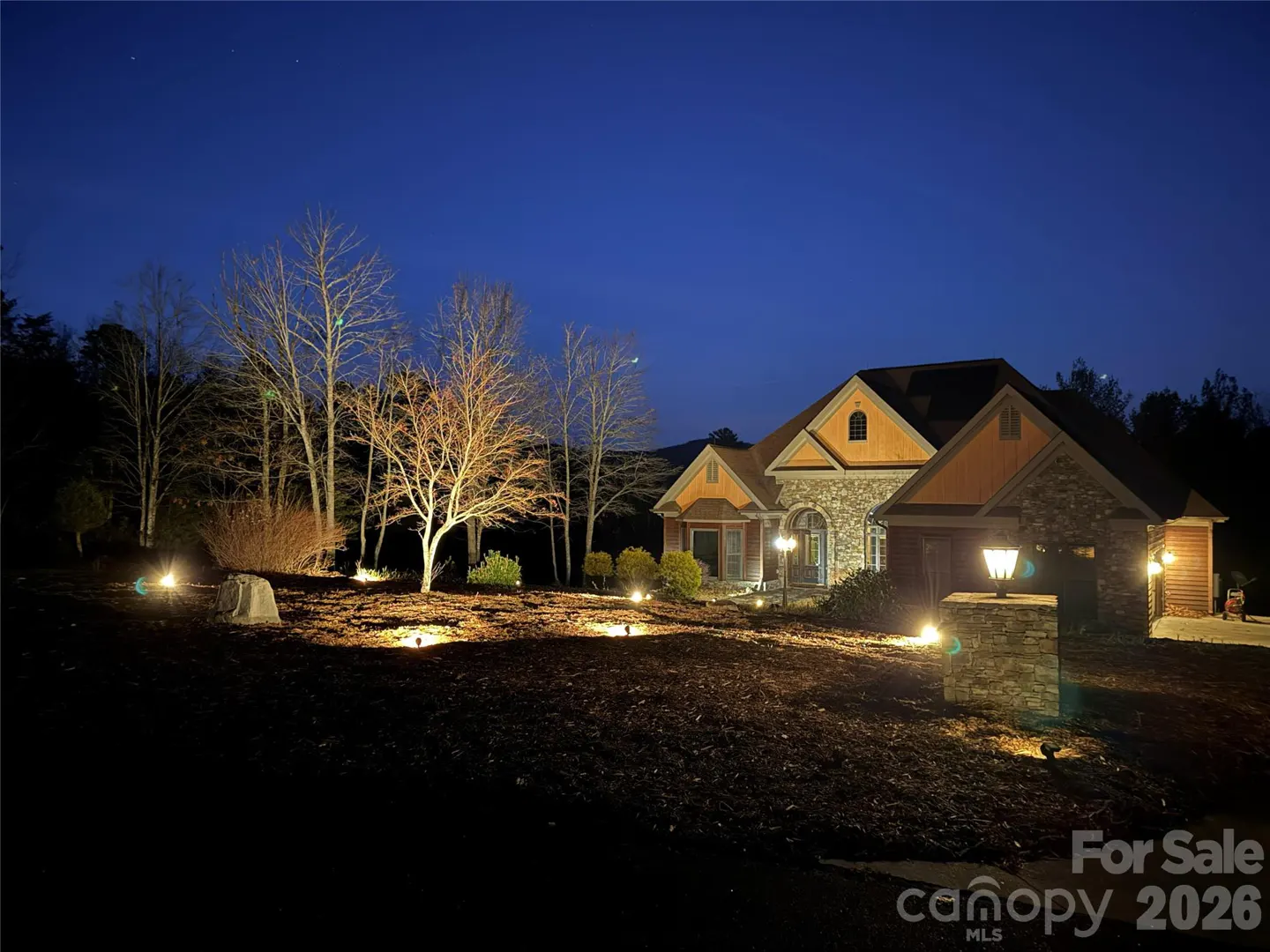 Night view of a stone and wood home with landscape lighting under a dark blue sky.