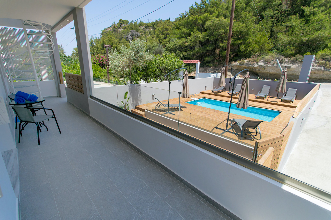 Balcony view of a blue pool with lounge chairs and umbrellas on a wooden deck, with green trees in the background. A table and chairs are on the balcony. [AI-generated text]