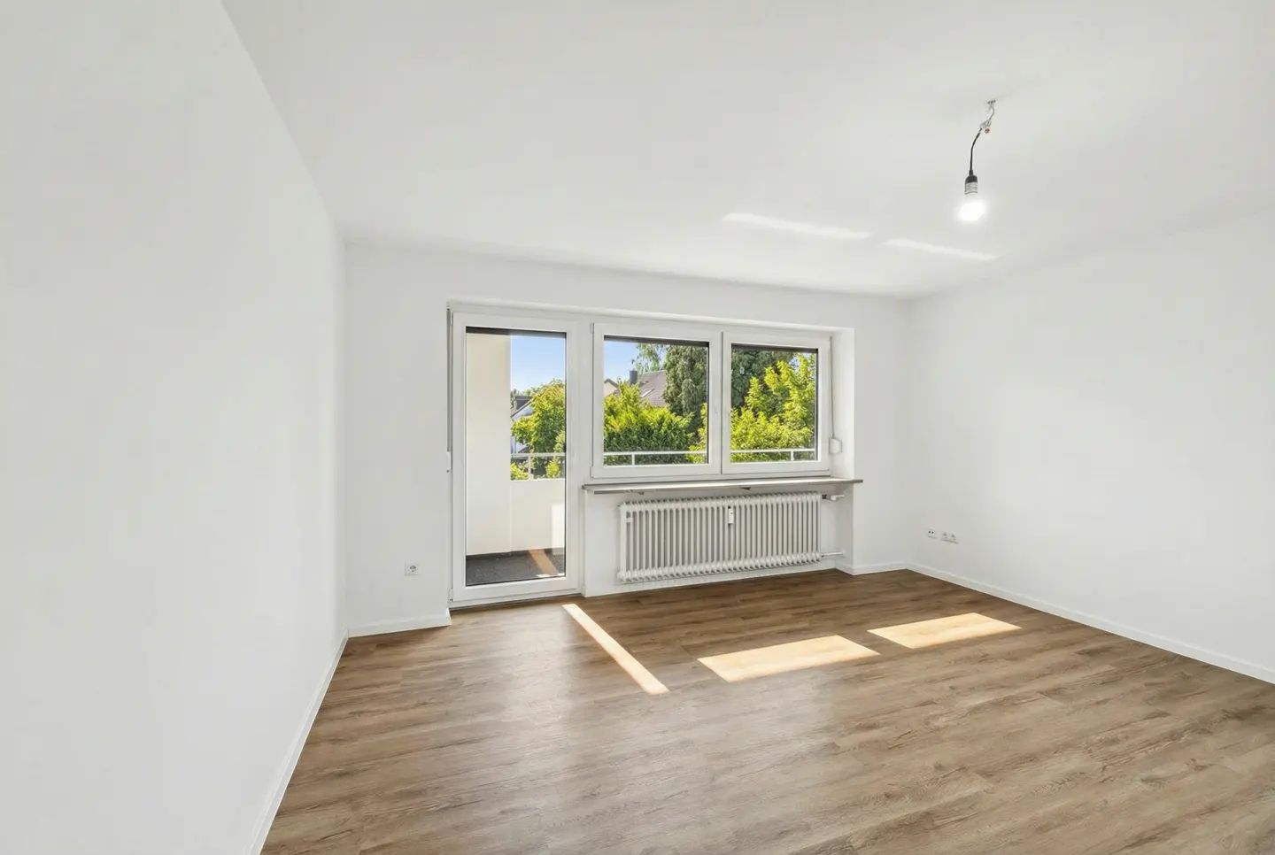 Bright, empty room with wood floors, white walls, and a window/door to a balcony with green trees outside.