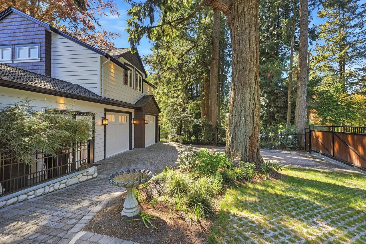 Exterior view of a two-story house with white siding, a gray roof, and a two-car garage, surrounded by trees and greenery.
