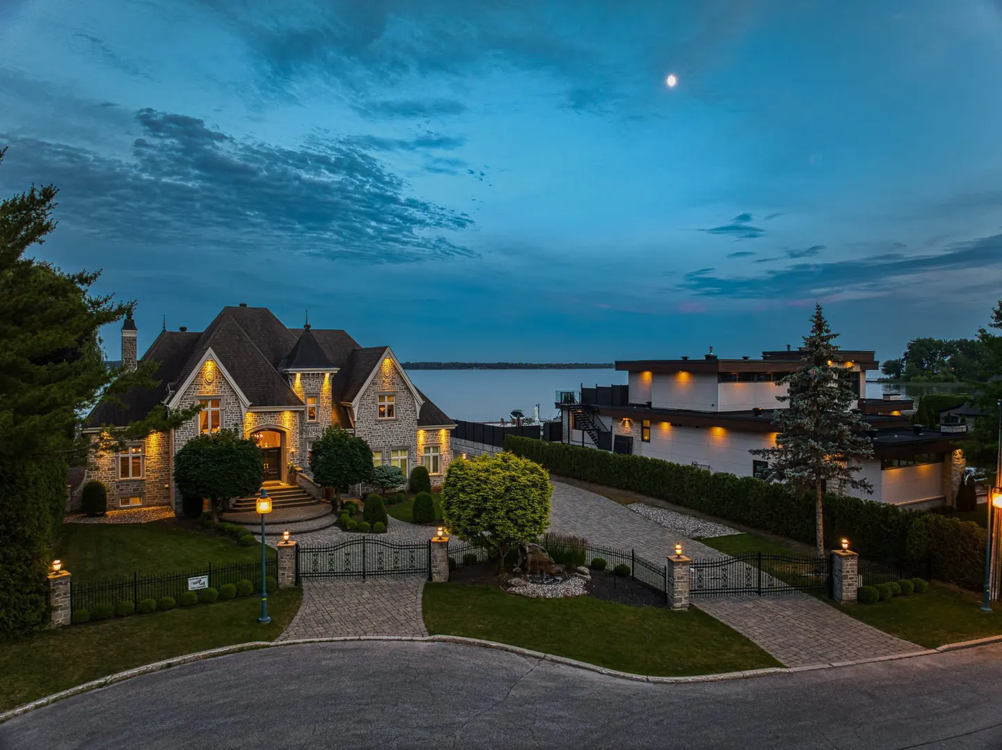 A large stone house with lights on at dusk, with a lake in the background.