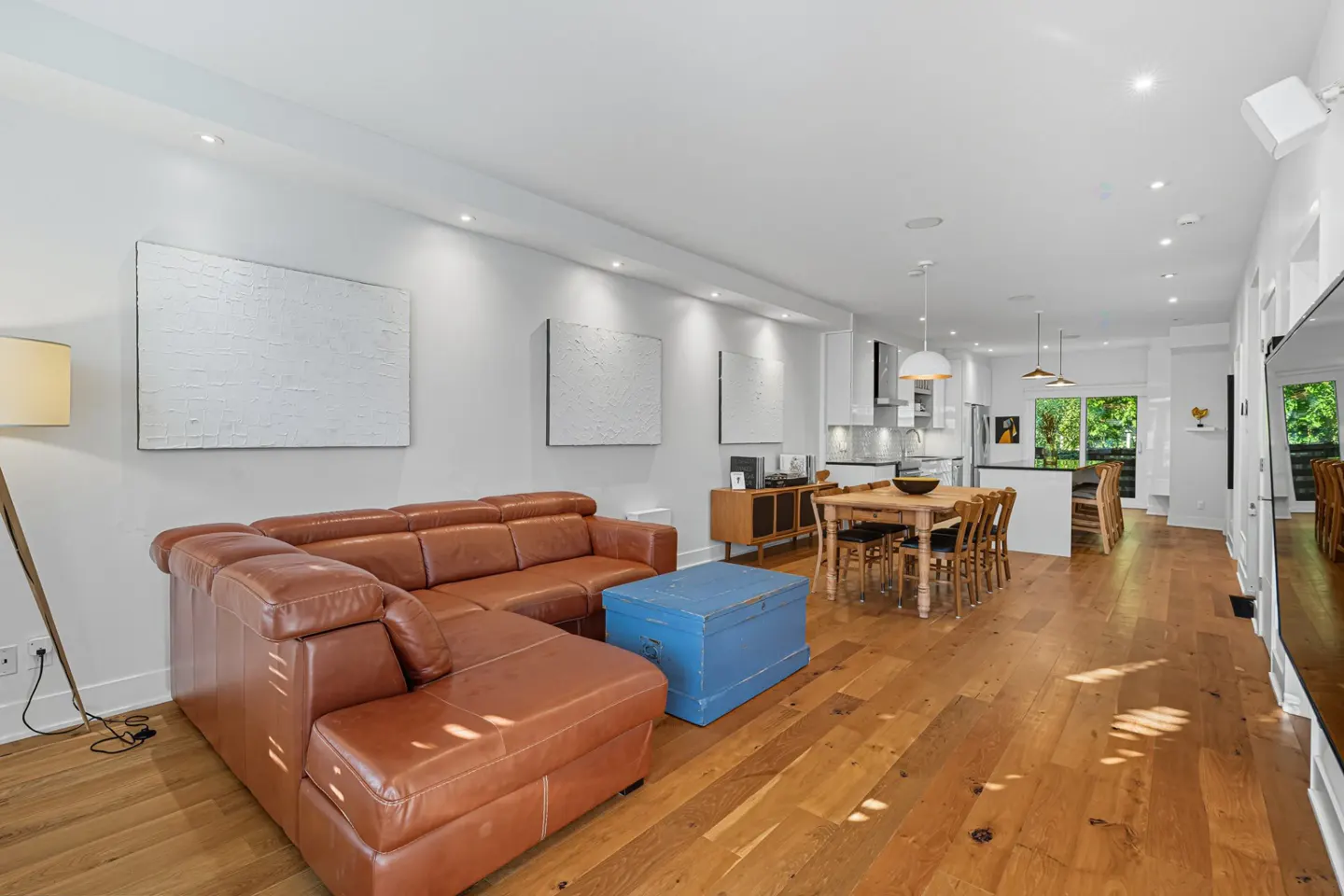 Open-concept living space with a brown leather sectional, blue chest, and wood floors. White walls feature abstract art. Kitchen in the background.