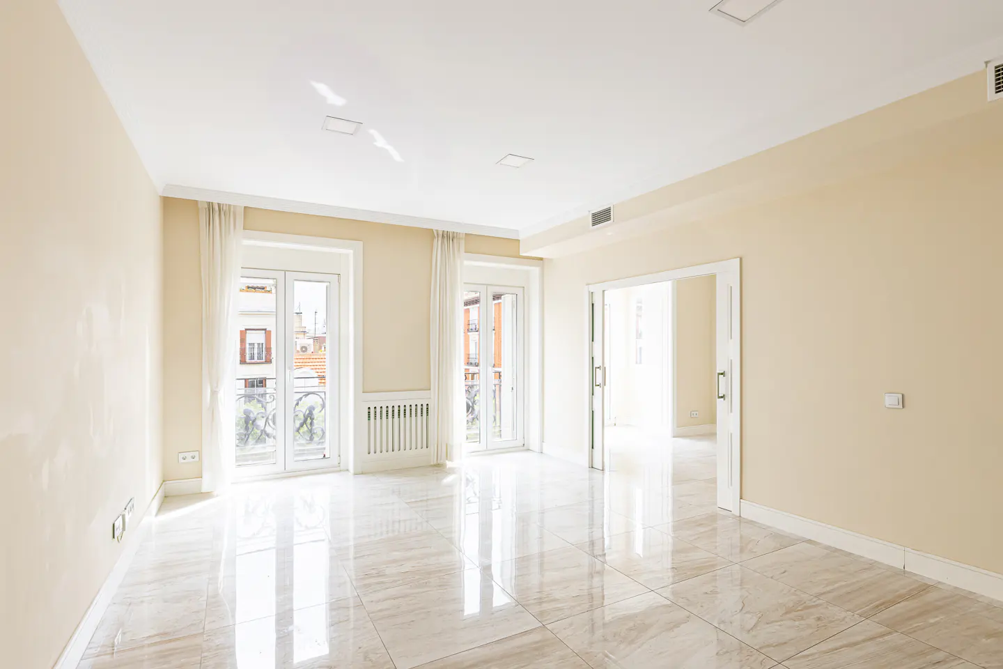 Bright, empty room with beige walls, marble floors, and white trim. Two sets of French doors lead to balconies.