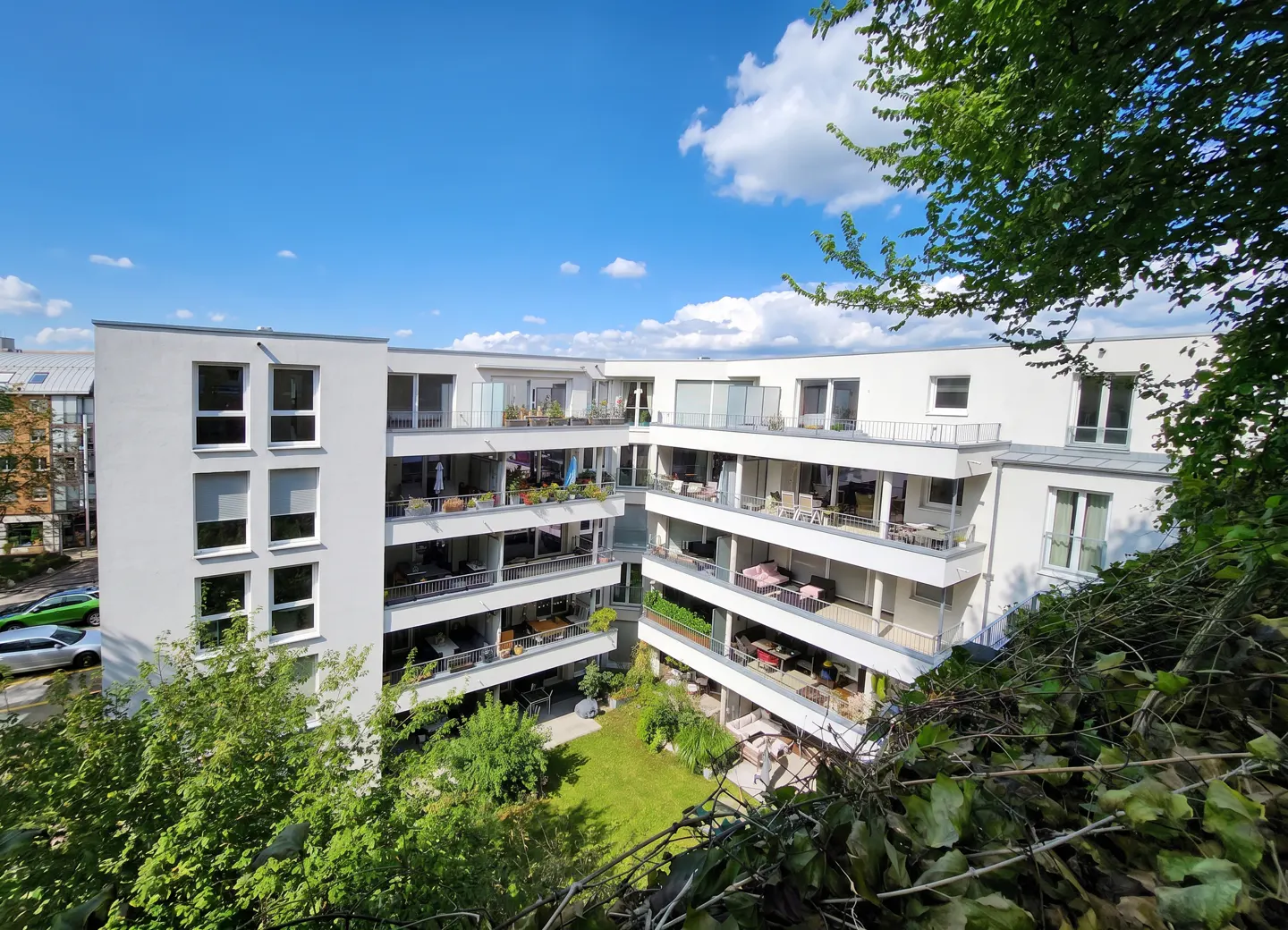 Exterior view of a modern white apartment building with balconies overlooking a green courtyard on a sunny day.