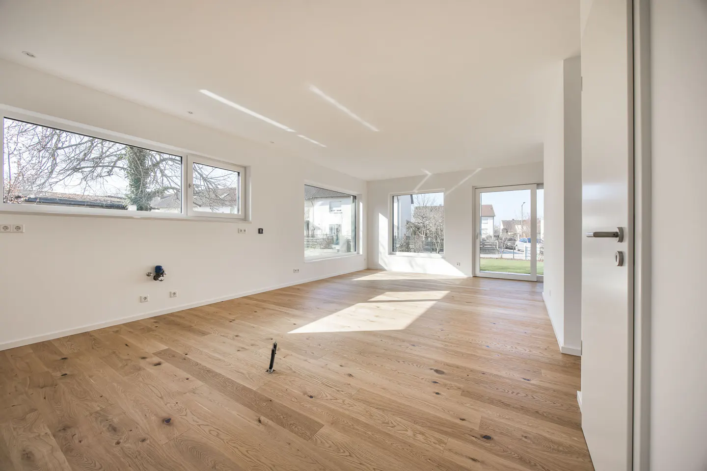 Bright, empty room with light wood floors, white walls, and large windows. Sunlight streams in, highlighting the open space. A white door is ajar.