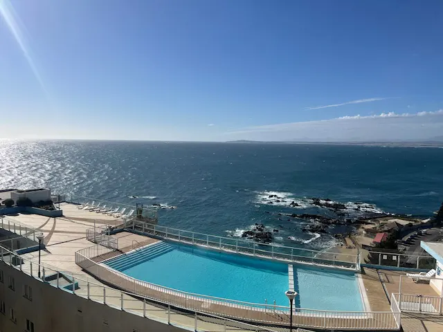 Ocean view with a blue pool in the foreground, white lounge chairs, and a clear blue sky.