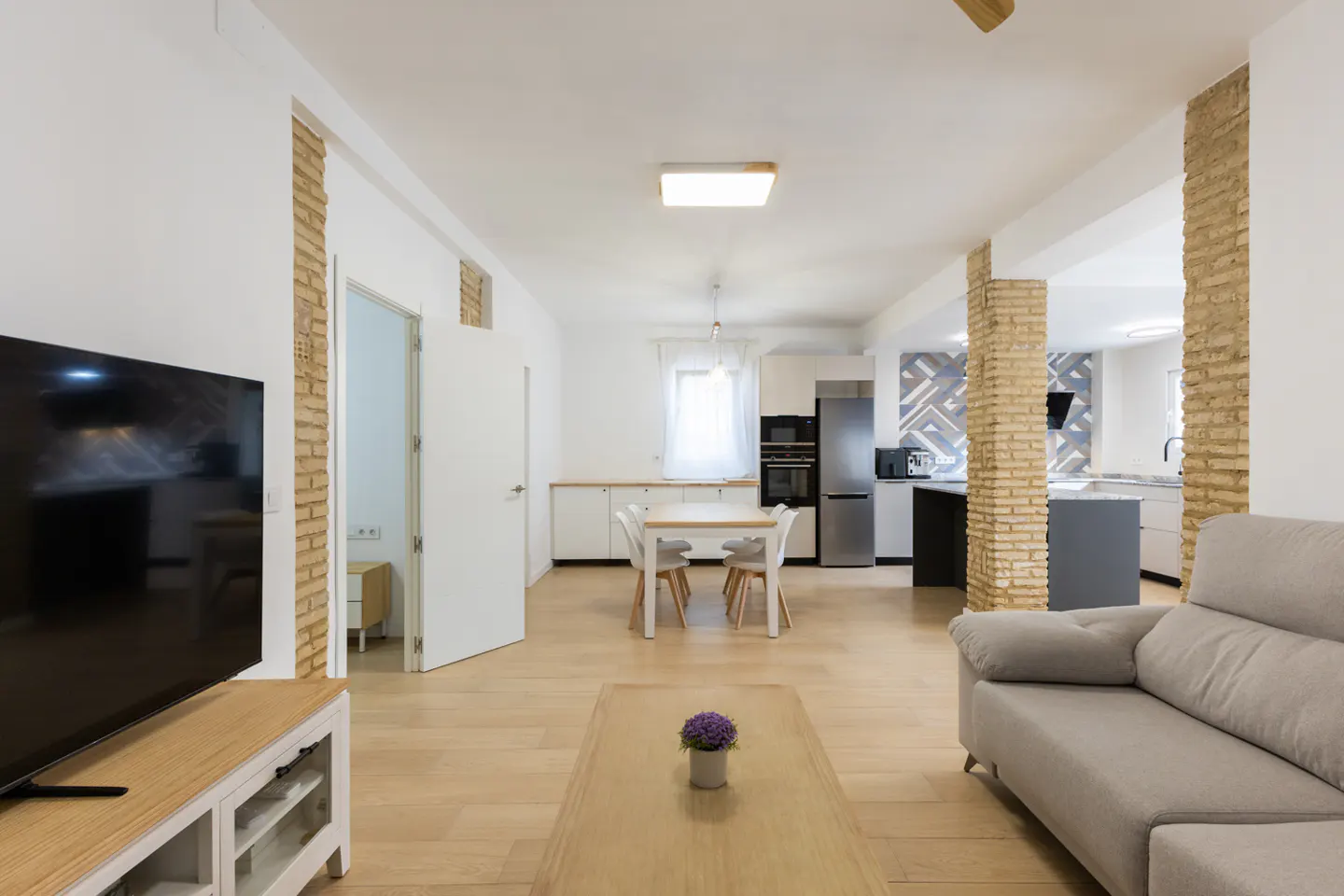 Bright, open-concept living space with wood floors, a gray sofa, and a TV. The kitchen features white cabinets and stainless steel appliances.