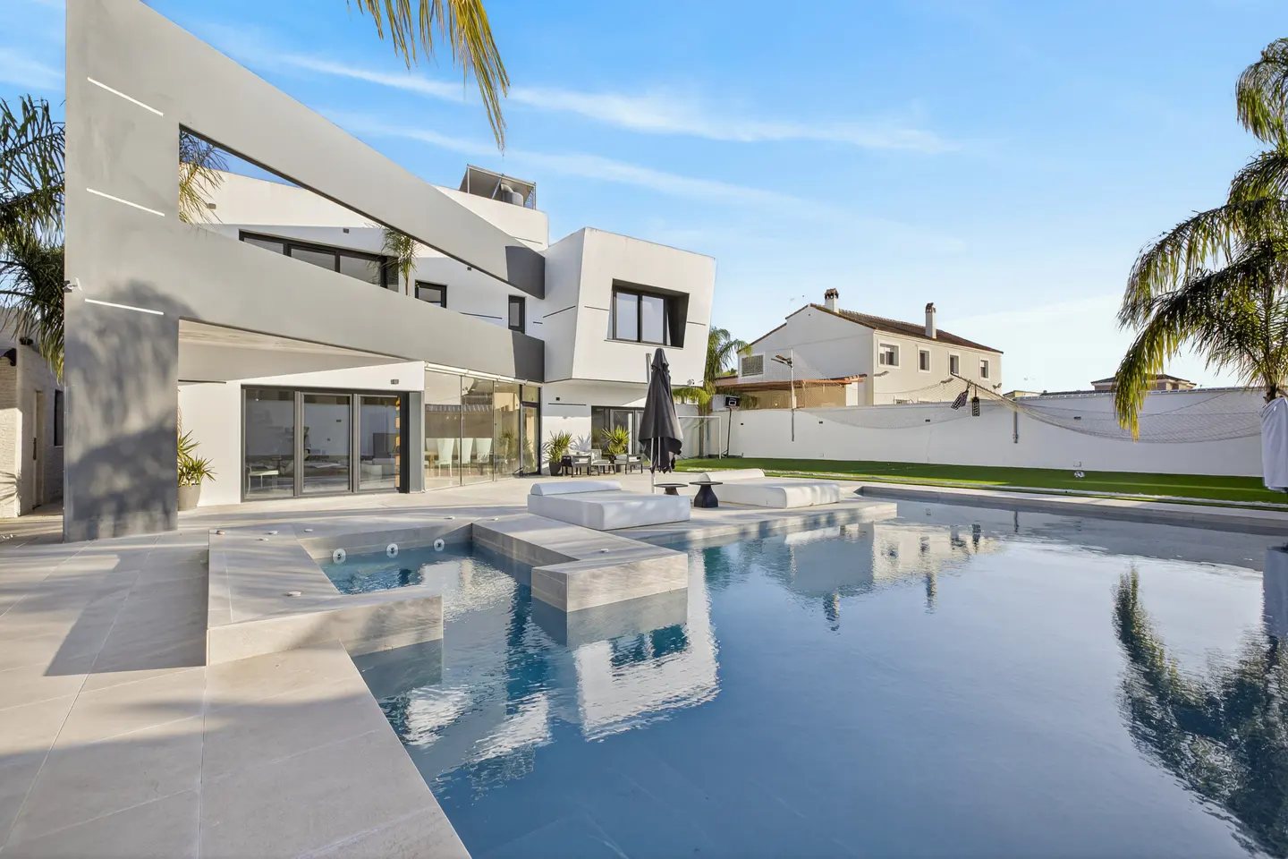 Modern white house with a pool. Lounge chairs and an umbrella sit on the patio. Blue sky.