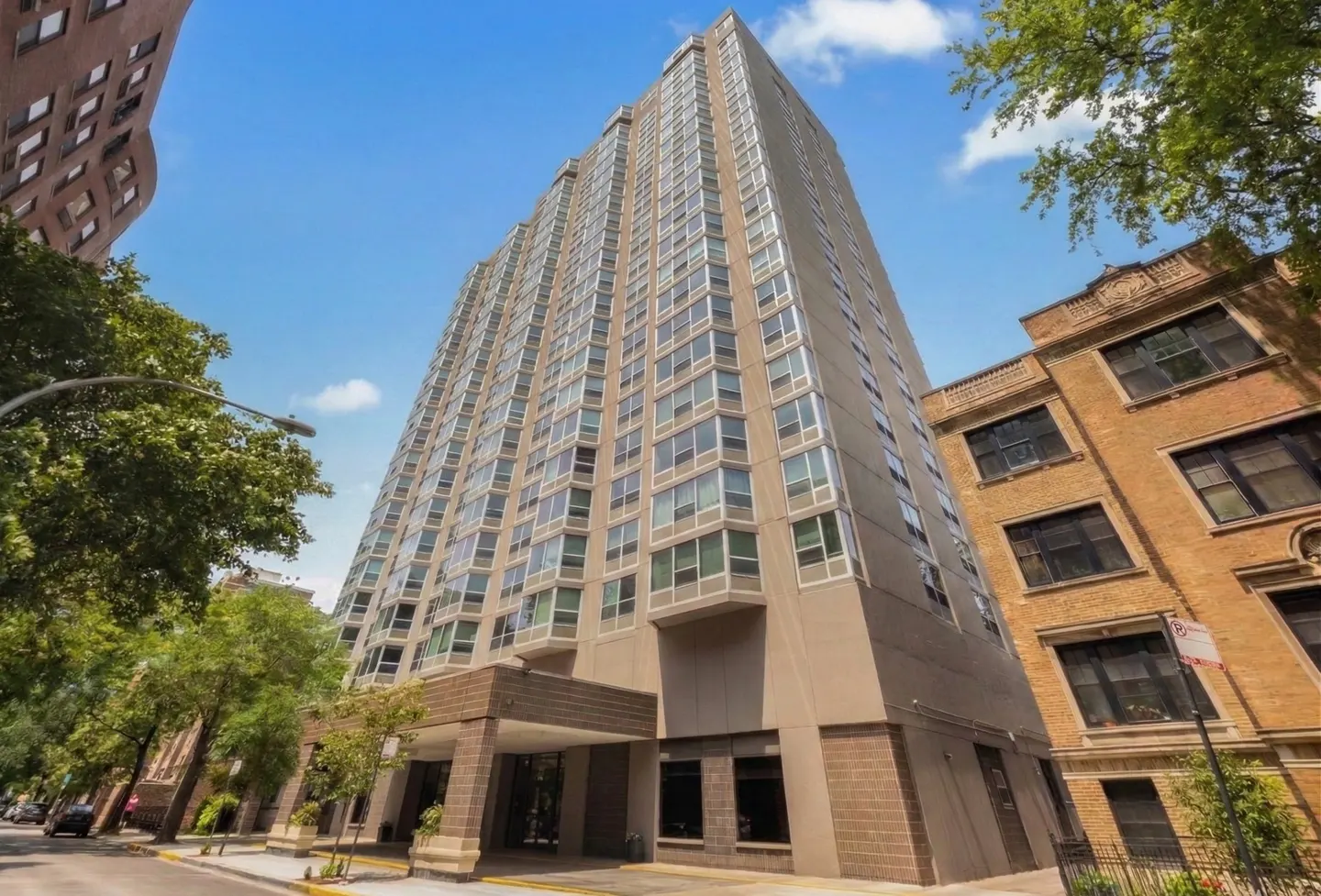 Exterior view of a tall, modern apartment building with many windows, set against a blue sky with trees nearby.