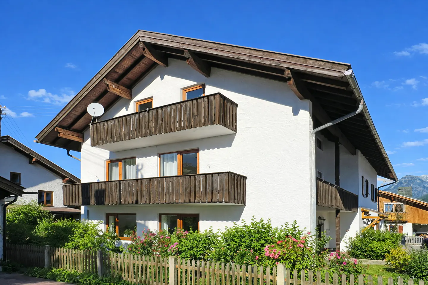 A two-story white house with dark wood balconies and a brown roof under a blue sky. A wooden fence and green bushes are in front.
