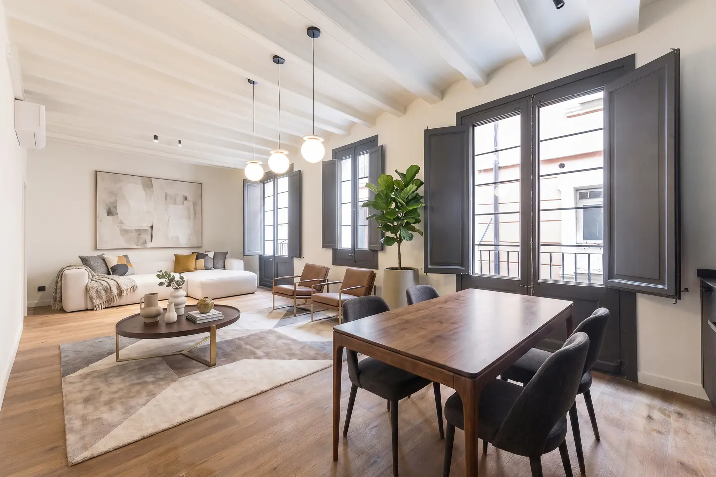 A bright living and dining area with a white sofa, wood table, and large windows with dark shutters.