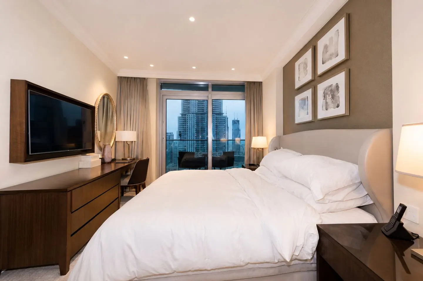 Bedroom with white linens, dark wood furniture, and a balcony view of city skyscrapers. Four framed pictures hang above the bed.