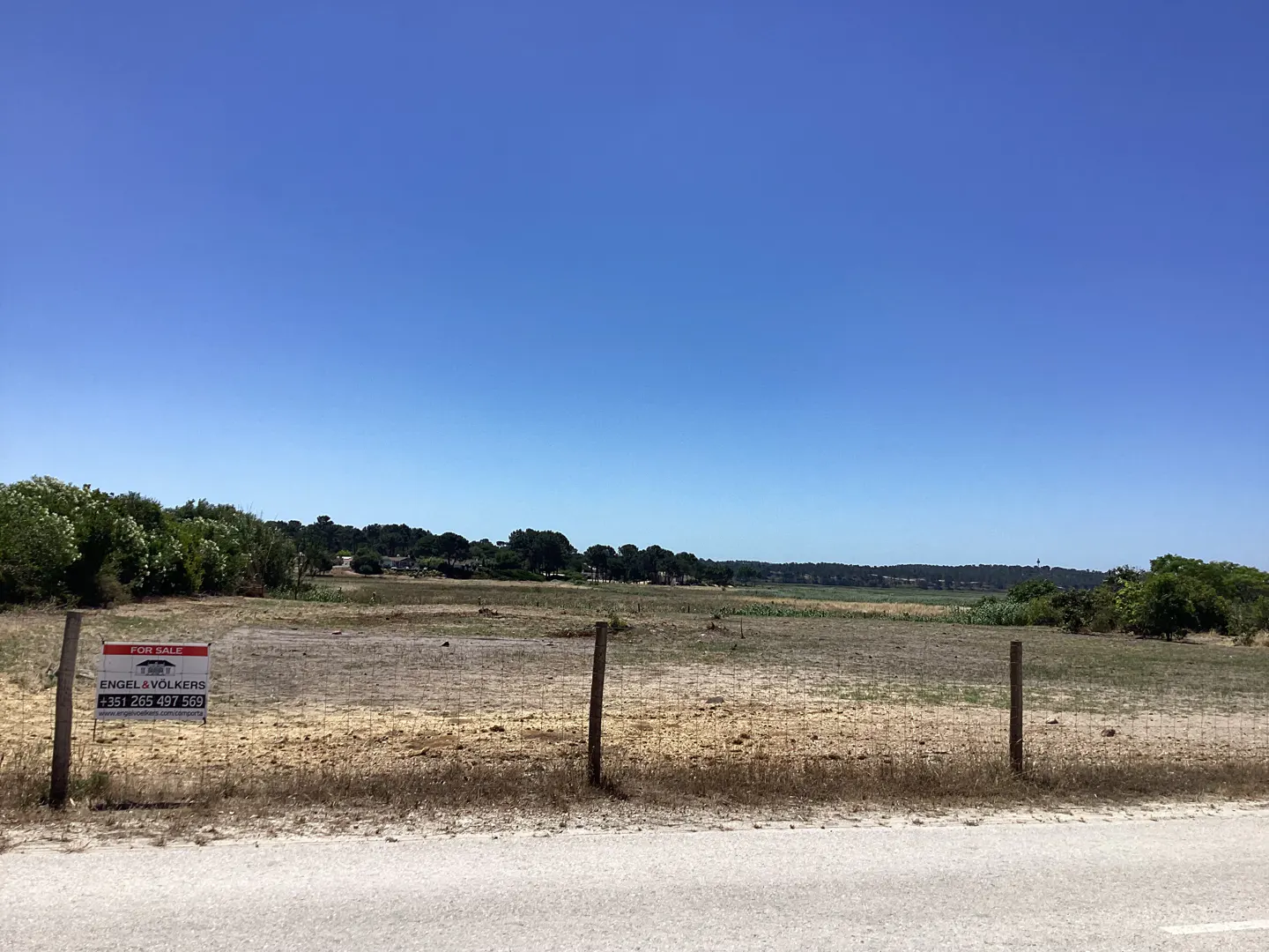 Vacant plot of land for sale with an Engel & Völkers sign, bordered by a wire fence under a clear blue sky.