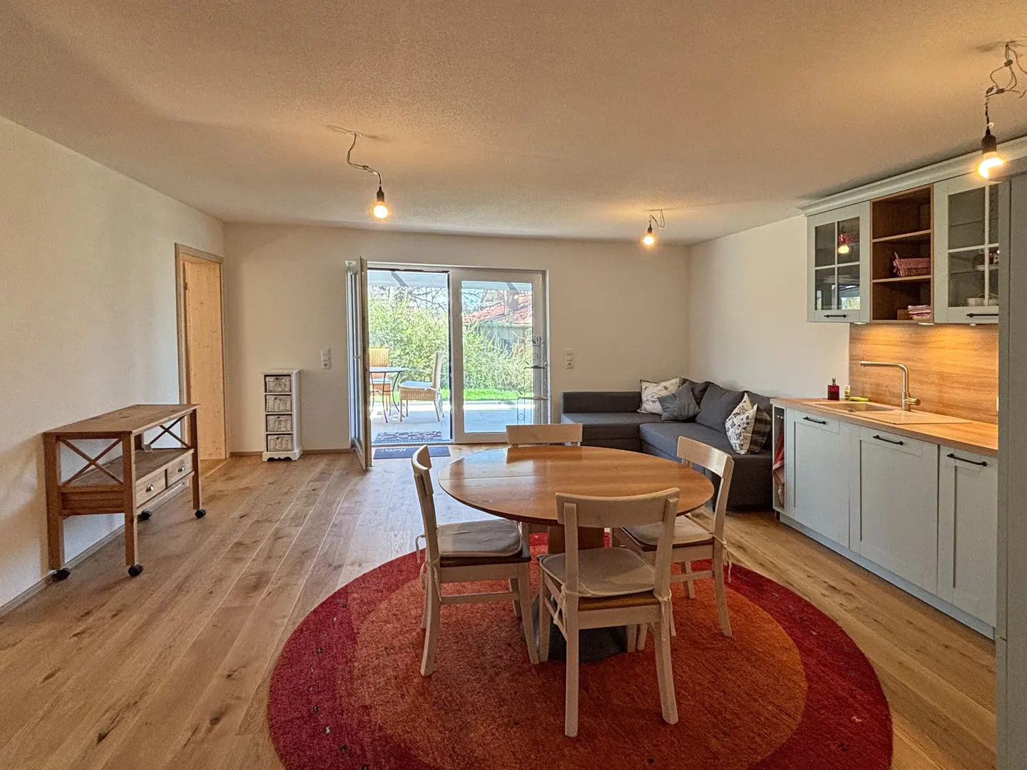 Open-plan living space with wood floors, a round table with chairs on a red rug, a gray sofa, and light blue kitchen cabinets.