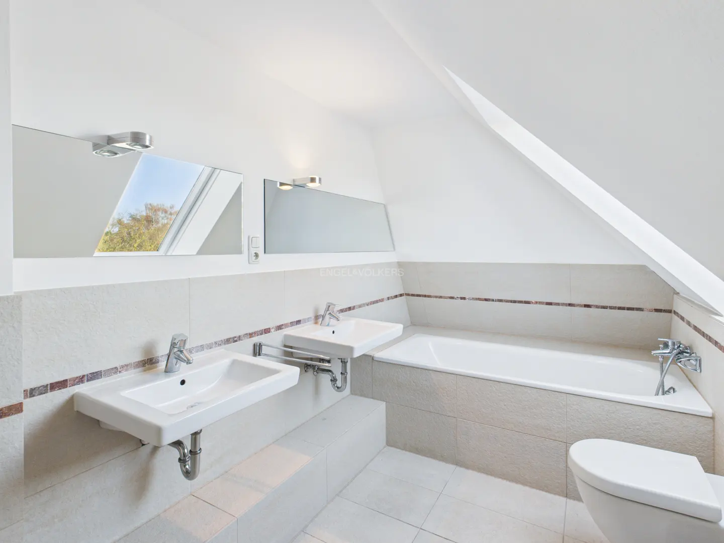 Bright bathroom with two sinks, a tub, and a toilet. Mirrors reflect a window. The room has an angled ceiling.