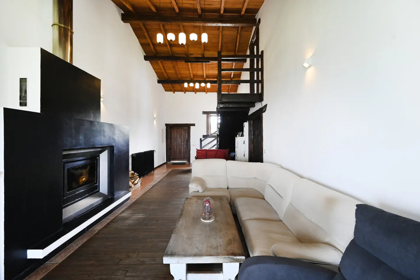 Living room with a black fireplace, wood floors, and a white sectional sofa. A wooden ceiling and staircase add rustic charm.