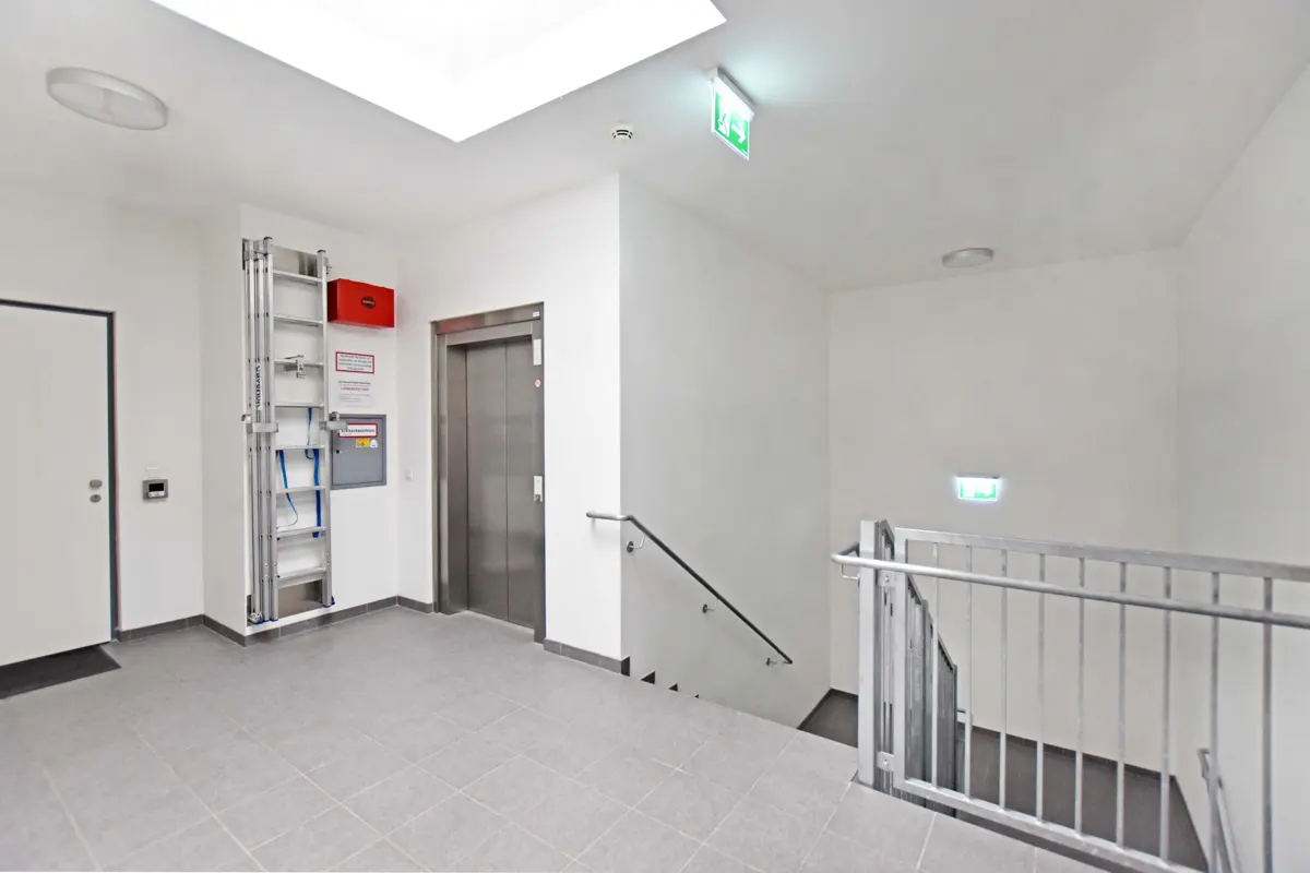 A bright, white hallway with a gray tiled floor, an elevator, stairs with metal railings, and a ladder.