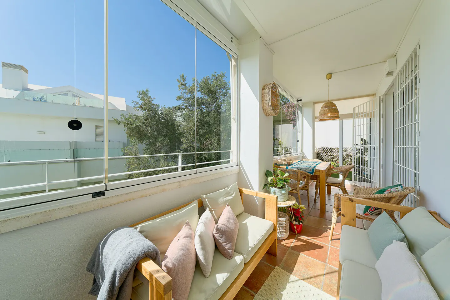 Bright sunroom with terracotta tile floor, white walls, and glass doors. Two wooden sofas with cushions face a dining table with wicker chairs.
