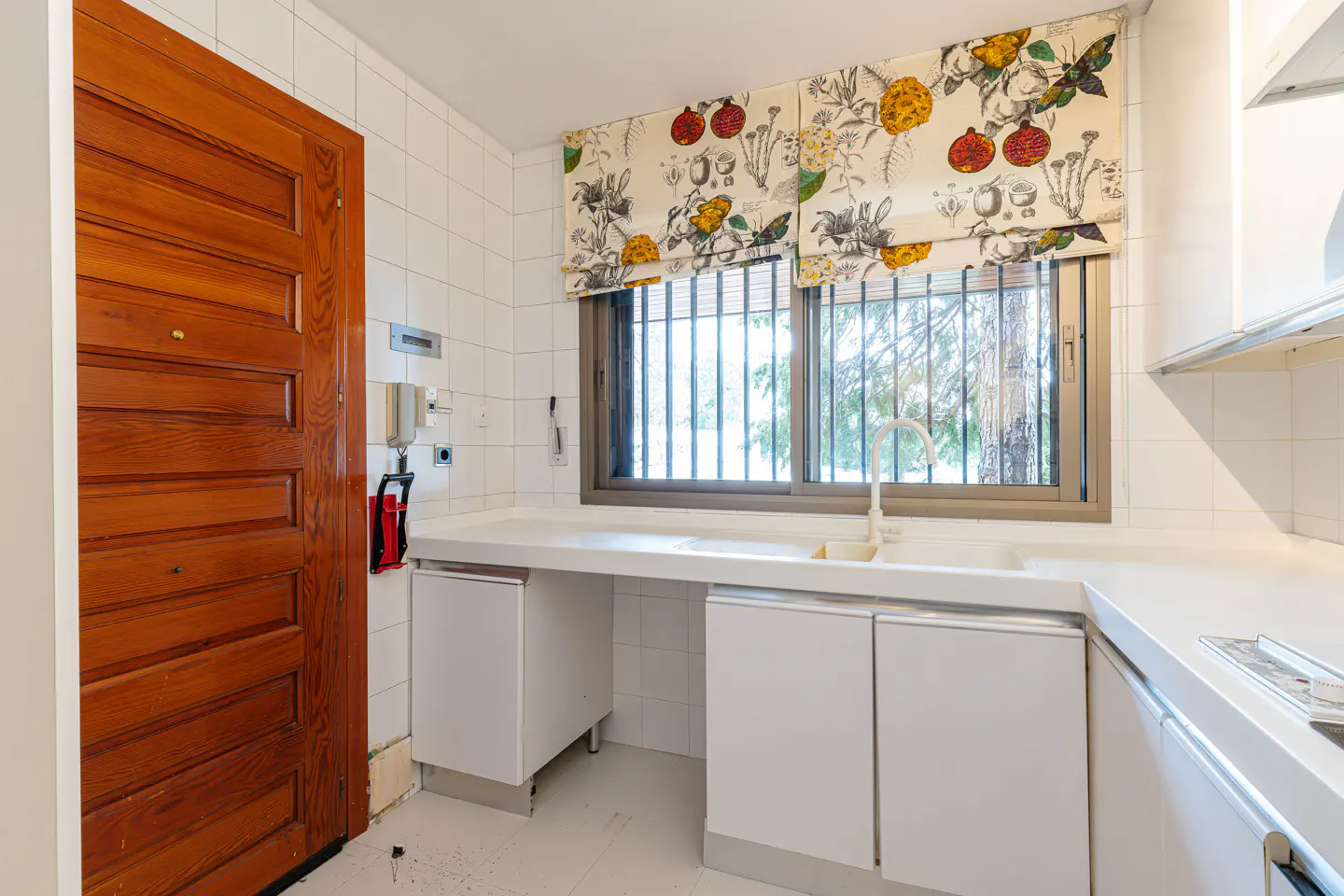A bright kitchen with white cabinets, a white countertop, and a window with floral print blinds. A wooden door is on the left.