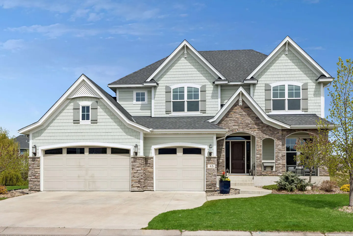 Two-story house with light green siding, stone accents, a gray roof, and a two-car garage on a sunny day.
