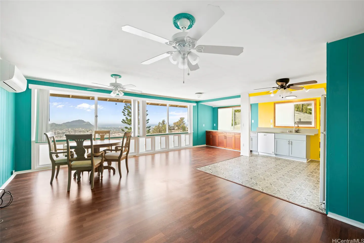 Bright, open-concept living space with hardwood floors, turquoise walls, and a dining table with a scenic view. Kitchen with yellow accents.