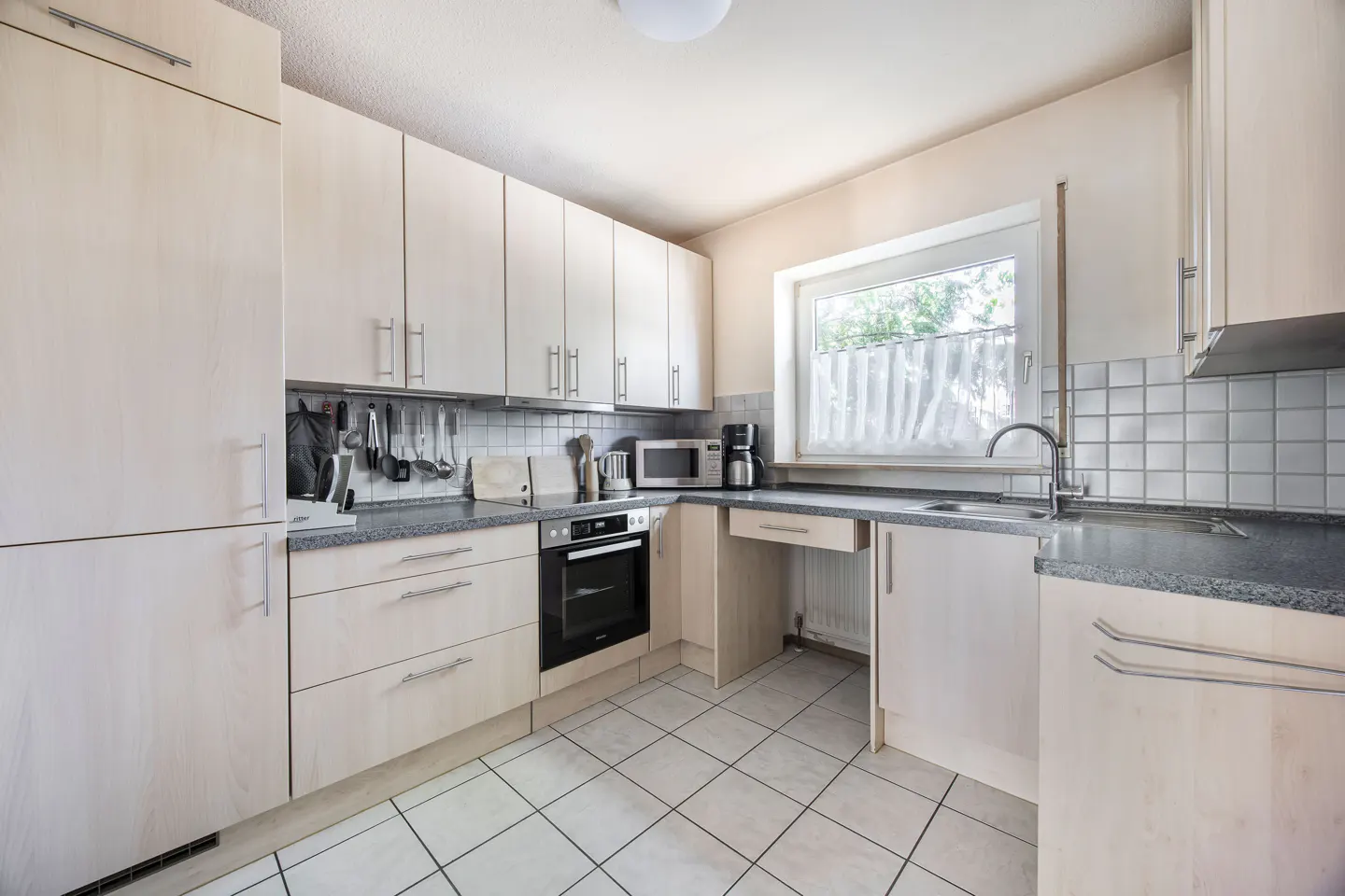 A bright kitchen with light wood cabinets, gray countertops, and white tiled floors. A window is above the sink.