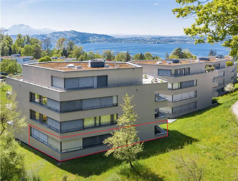 Modern apartment building with a flat roof and green lawn, overlooking a lake and mountains. A red line highlights the ground floor.