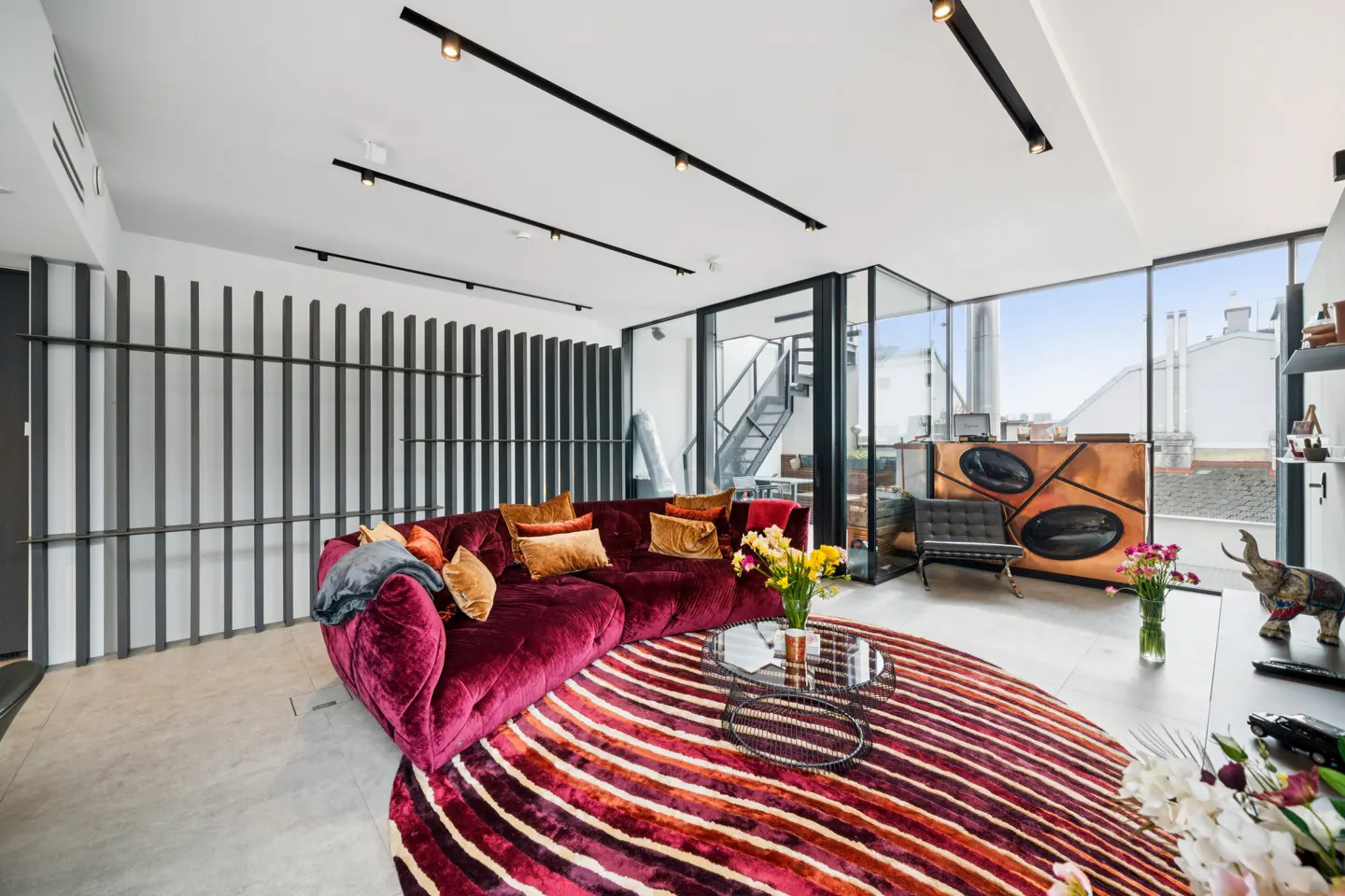 Bright living room with a curved, burgundy velvet sofa, gold pillows, and a striped rug. Track lighting and floor-to-ceiling windows.
