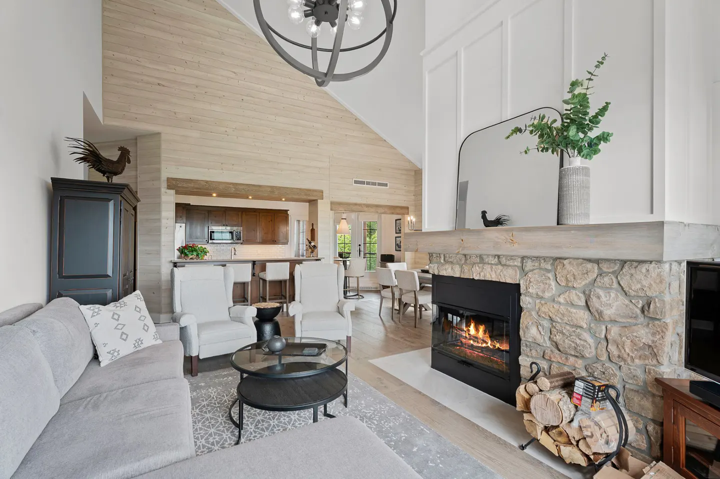A bright, open living space with a stone fireplace, gray sofa, and kitchen in the background.