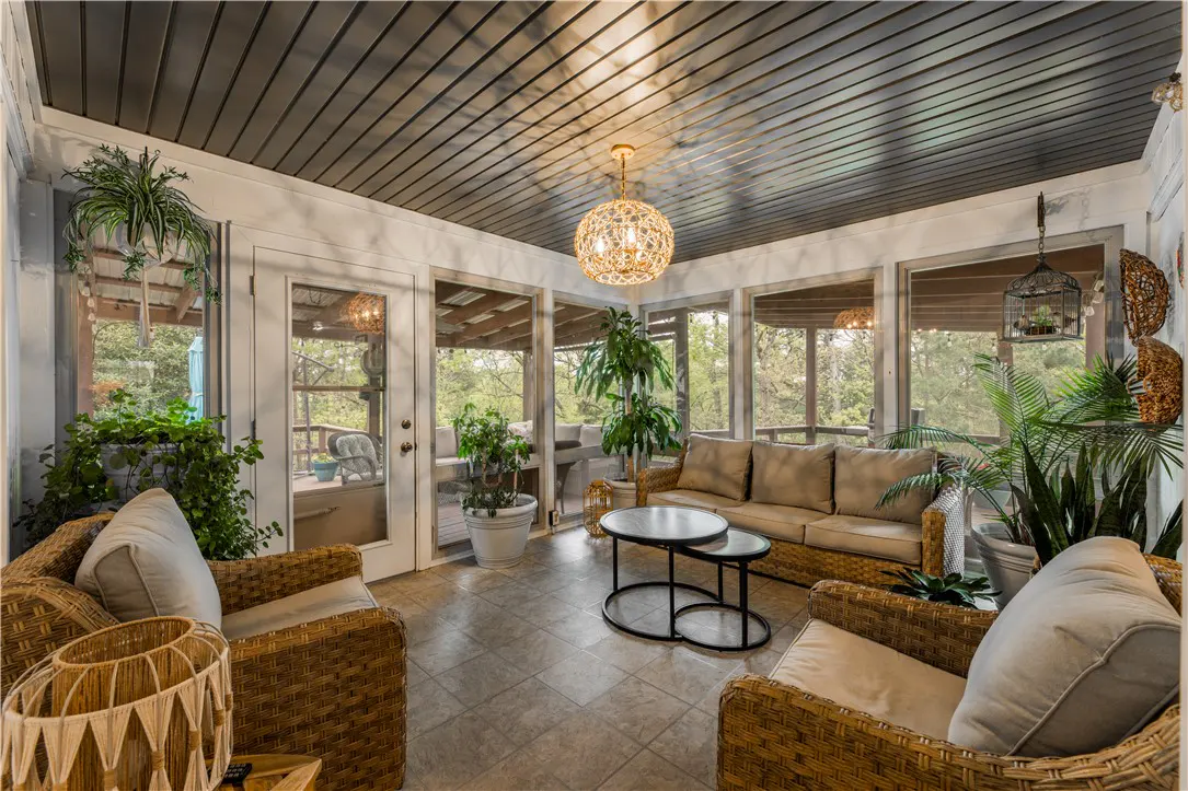 Sunroom with wicker furniture, beige sofa, and plants. A decorative light fixture hangs from the gray plank ceiling. Windows overlook a wooded area.