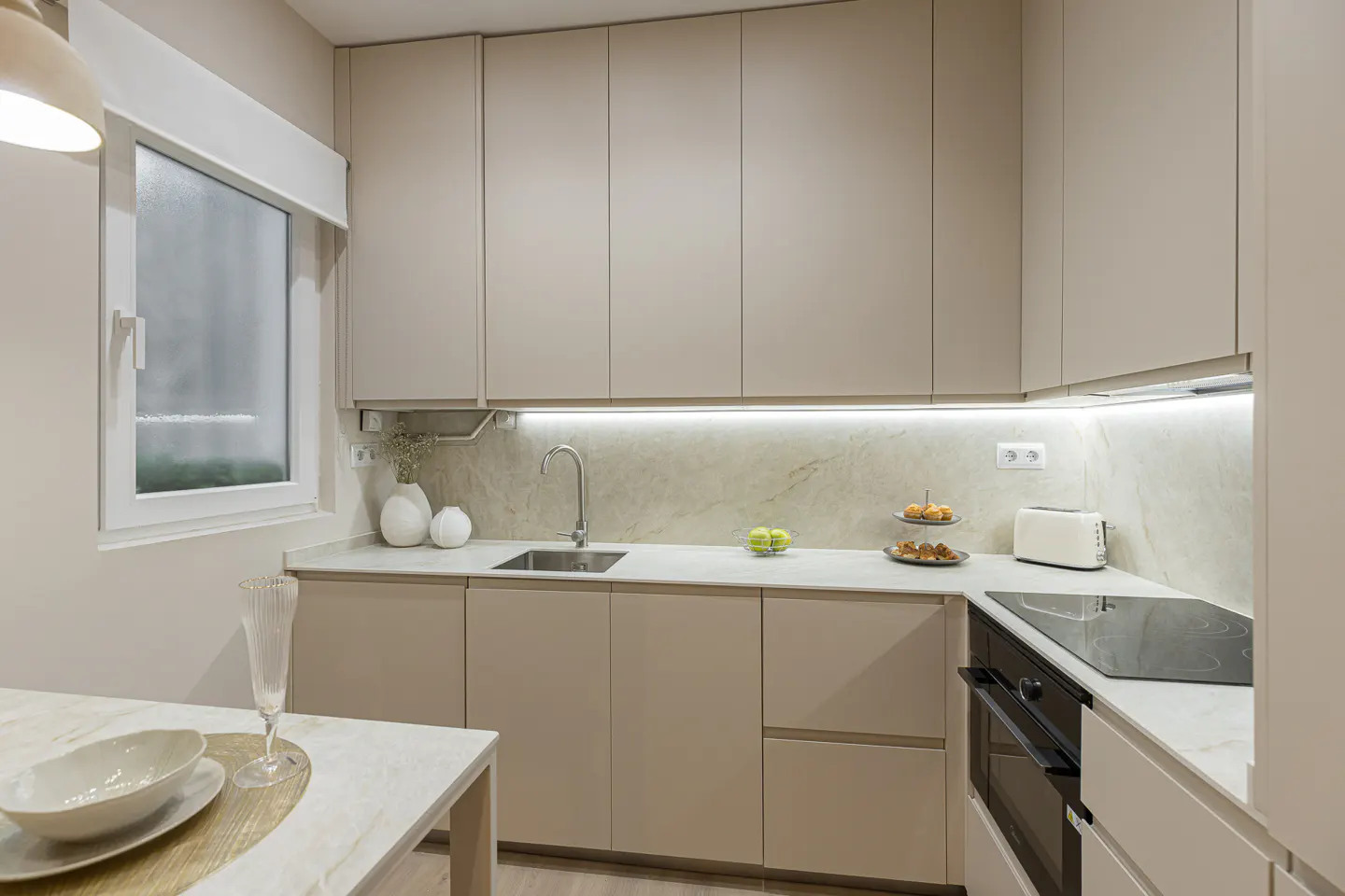Modern kitchen with beige cabinets, white countertops, and stainless steel sink. A table with dishes is in the foreground.